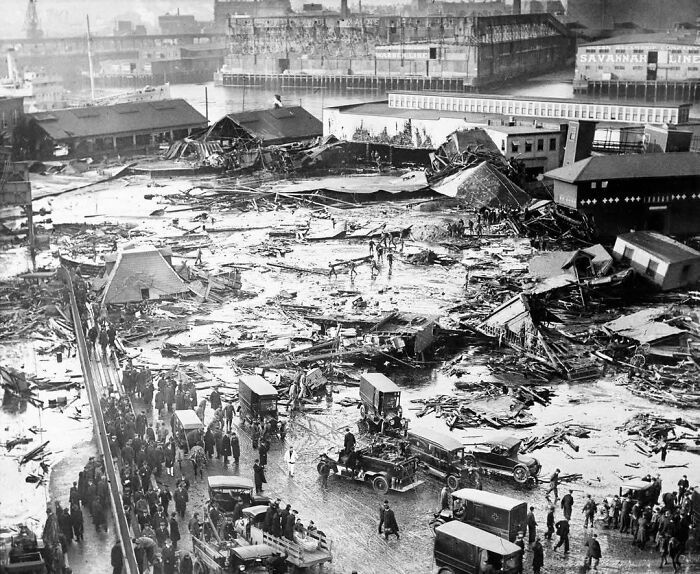 Crowd and vehicles gather around extensive destruction at a waterfront disaster site in a fascinating historical photo.