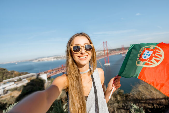 Young woman in sunglasses holding Portuguese flag outdoors with bridge in background, illustrating European netizens correcting common country facts.