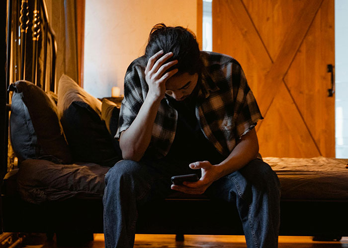 Man sitting on bed looking distressed, holding phone, reflecting on wife cheating and her mind-controlled excuse.