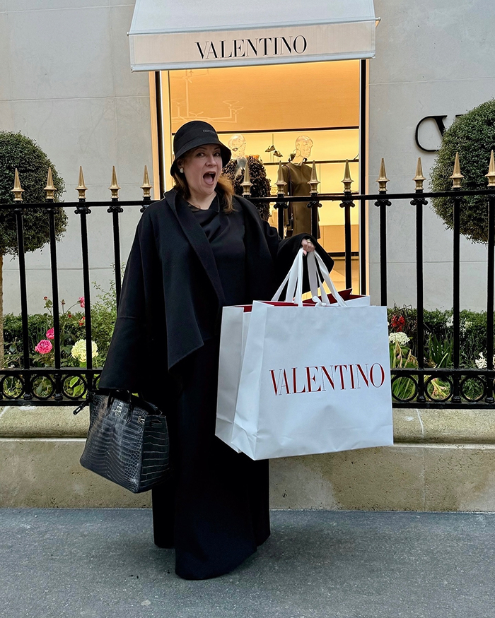 Jennifer Tilly holding Valentino shopping bags outside a store, dressed in black with a hat and handbag.