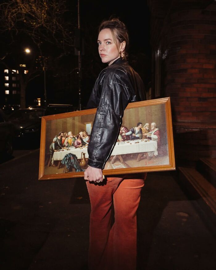 Young woman in a leather jacket holding a framed painting on a dimly lit street in candid street photography style.