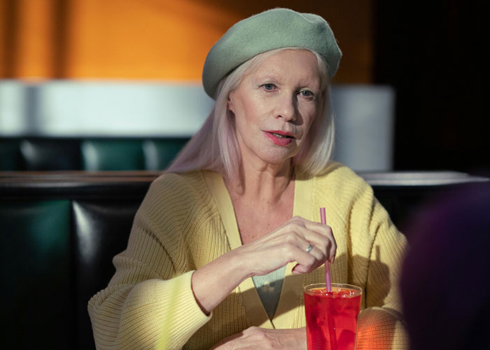 Older woman in a yellow sweater and beret sipping a red drink during an awkward date in a dimly lit diner setting.