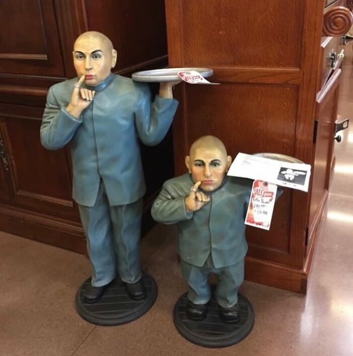 Two secondhand statues of a man in blue suits holding trays and making a shushing gesture inside a store.