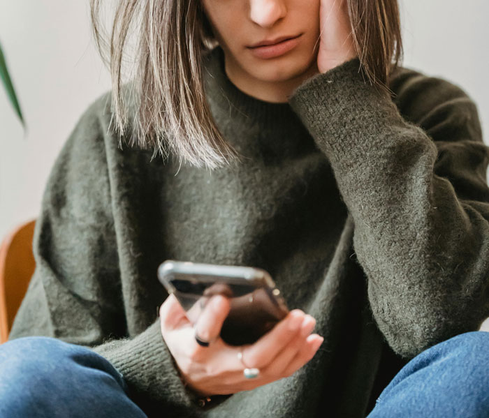 Young woman facing loneliness, looking troubled while holding a smartphone, wearing a green sweater and sitting indoors.