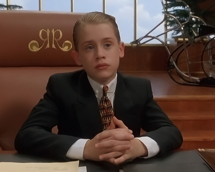 90s child star wearing a suit and patterned tie, sitting in a leather chair with a serious expression indoors.