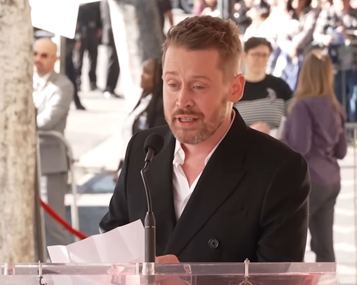 Beloved 90s child star at 44 speaking at an outdoor event, wearing a black blazer and white shirt.