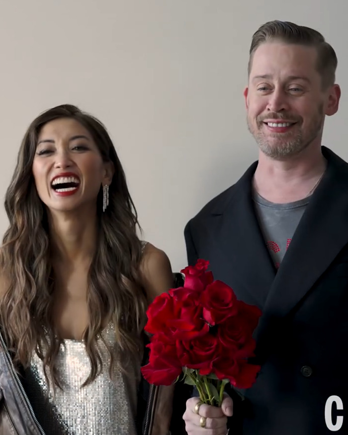 Man holding a bouquet of red roses beside a smiling woman wearing a silver dress in a casual indoor setting.
