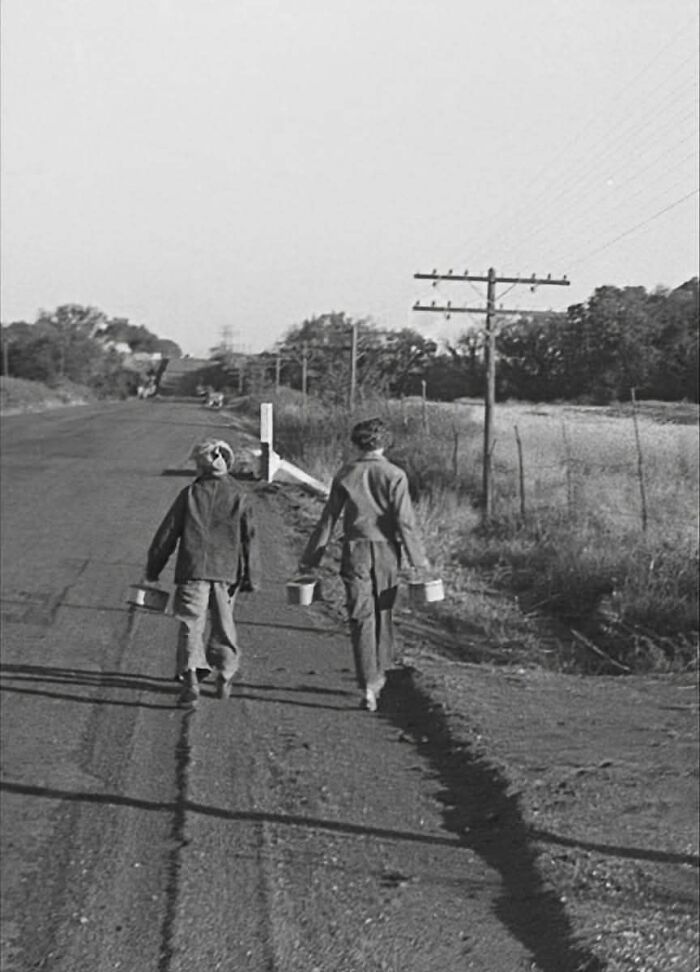 Two children walking along a rural road carrying buckets in a fascinating historical photo from the past.