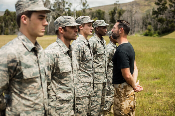 A group of soldiers in uniform standing in a line facing a man in civilian clothes, showing commitment to a lie.