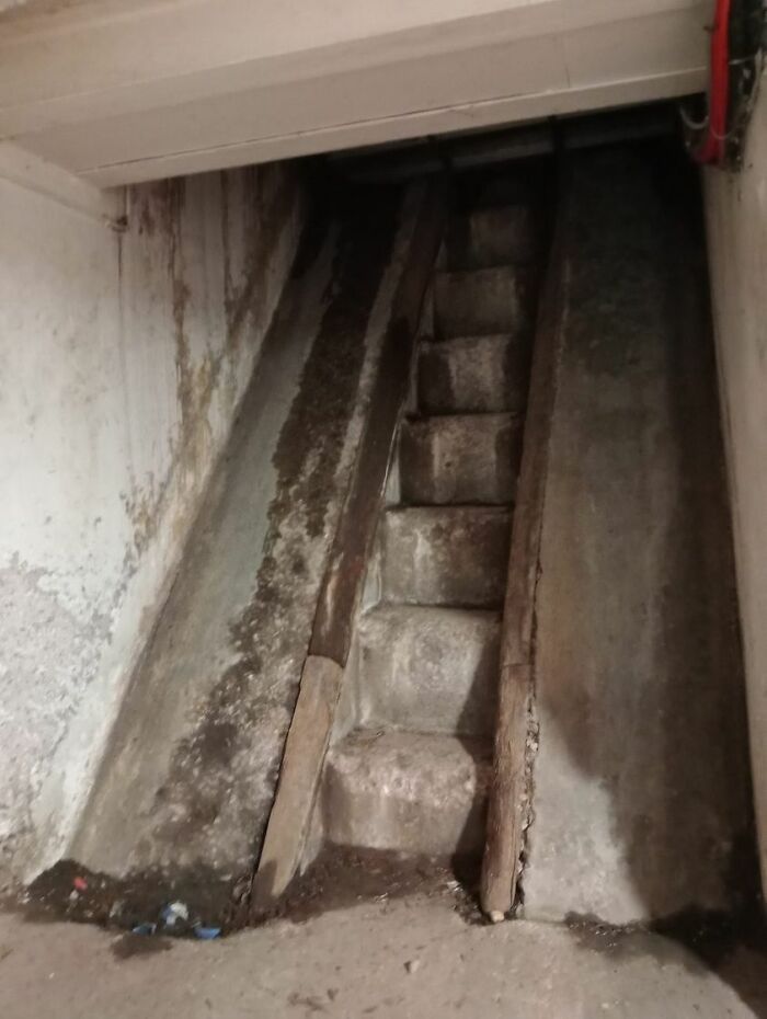 Steep and narrow death stairs with uneven concrete steps and wooden edges in a dimly lit, worn-down setting.