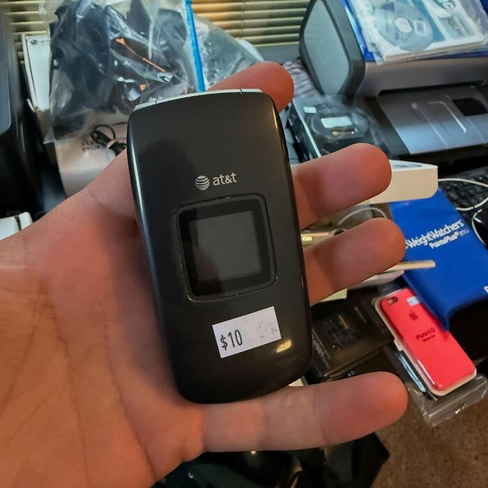 Hand holding a secondhand AT&T flip phone with a $10 price tag among other used electronics and items.