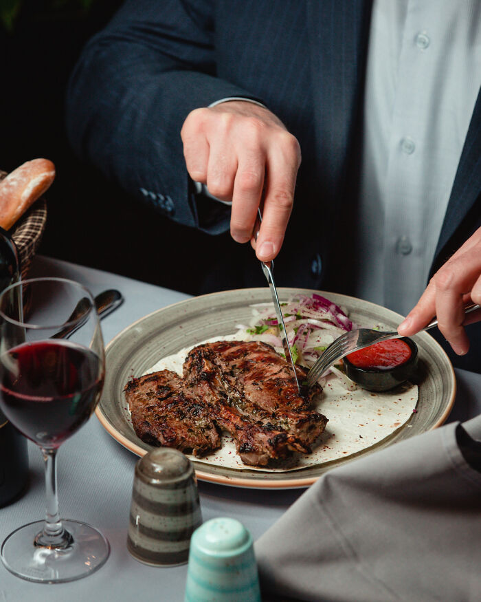 Man in a suit cutting steak on a plate with sauce and wine glass, illustrating out-of-touch rich people flaunting wealth.