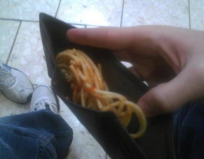 Hand holding an open wallet filled with cooked spaghetti noodles on a tiled floor, a cursed food picture.