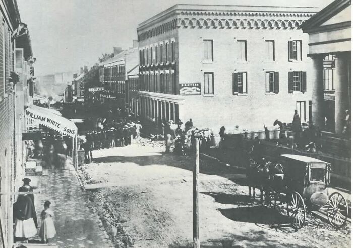 Black and white historical photo of a busy street with horse-drawn carriages and people, showcasing life in the past.
