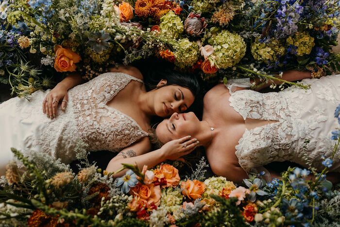Two brides in white lace dresses lying among colorful flowers in a stunning unforgettable wedding photo.