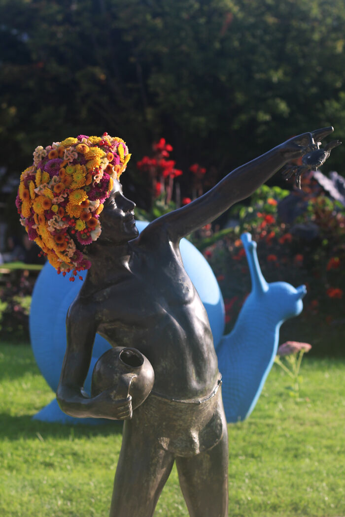 Bronze city statue dressed in vibrant flowers on its head, holding a jug, with a large blue snail sculpture in the background.