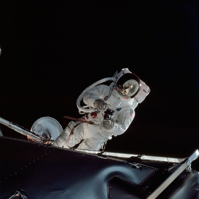 Astronaut in a white space suit taking photos during an Apollo mission spacewalk outside the spacecraft against a black space background.