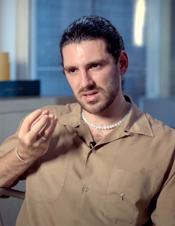 Young man in a brown shirt and pearl necklace, speaking seriously in an indoor interview about anti-vax mom controversy. Young man in a brown shirt and pearl necklace, speaking seriously in an indoor interview about anti-vax mom controversy.
