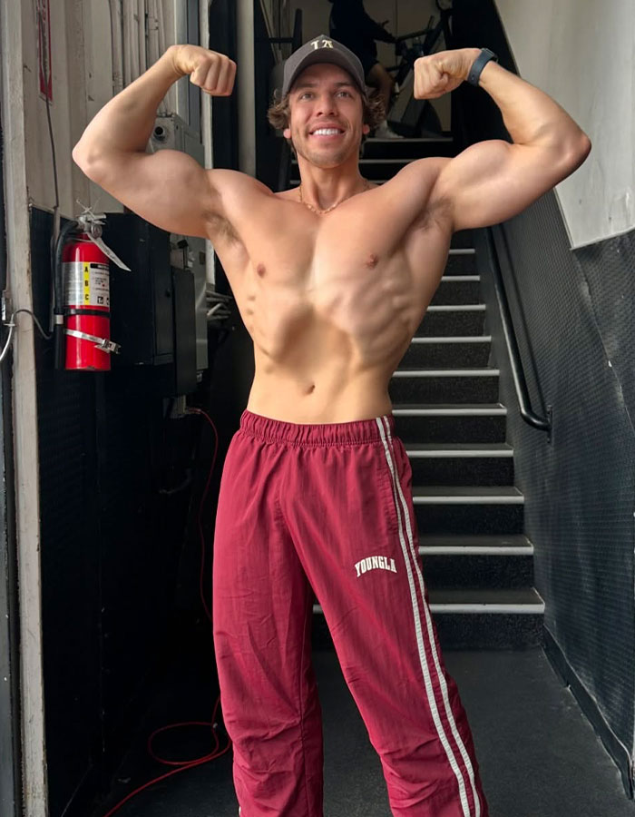 Muscular young man resembling Arnold Schwarzenegger flexing arms shirtless in casual pants near staircase indoors. Muscular young man resembling Arnold Schwarzenegger flexing arms shirtless in casual pants near staircase indoors.