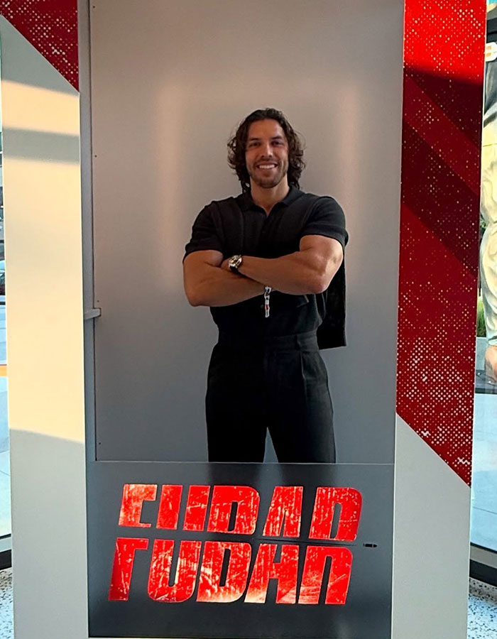 Man resembling Arnold Schwarzenegger posing with arms crossed at movie premiere event with bold red text below. Man resembling Arnold Schwarzenegger posing with arms crossed at movie premiere event with bold red text below.