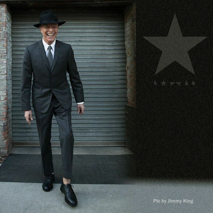 Famous person wearing a black suit and hat, smiling and walking outside near a metal garage door.