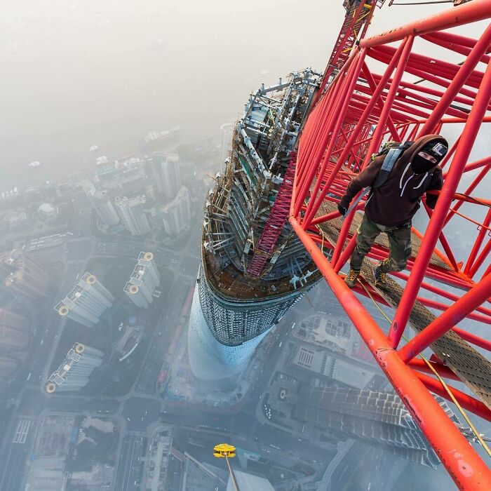 Person climbing a red crane on a famous landmark skyscraper showing an unexpected angle and cityscape below.