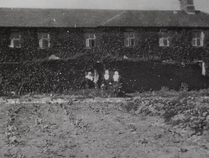 Black and white photo of a Catholic home for unwed mothers with a barren foreground and three people standing outside. Black and white photo of a Catholic home for unwed mothers with a barren foreground and three people standing outside.