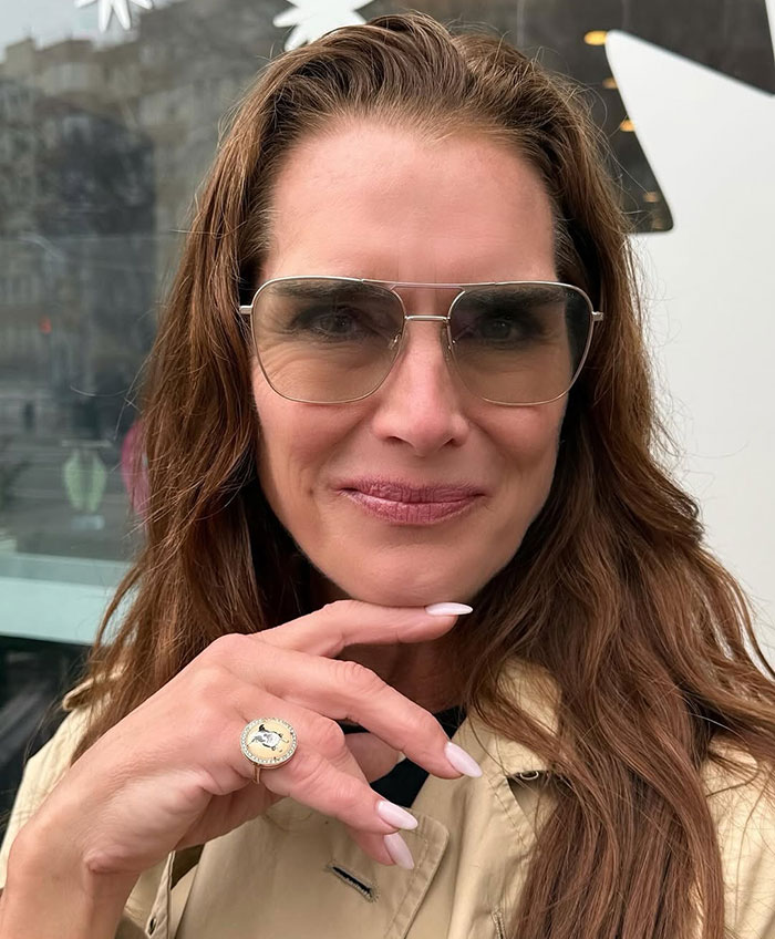 Brooke Shields wearing sunglasses and beige coat outdoors, linked to podcast interview about Meghan Markle controversy.