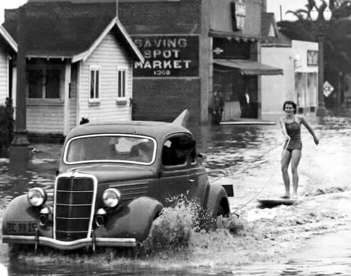 Vintage car driving through floodwaters while a woman water skis behind in a historic black and white photo.