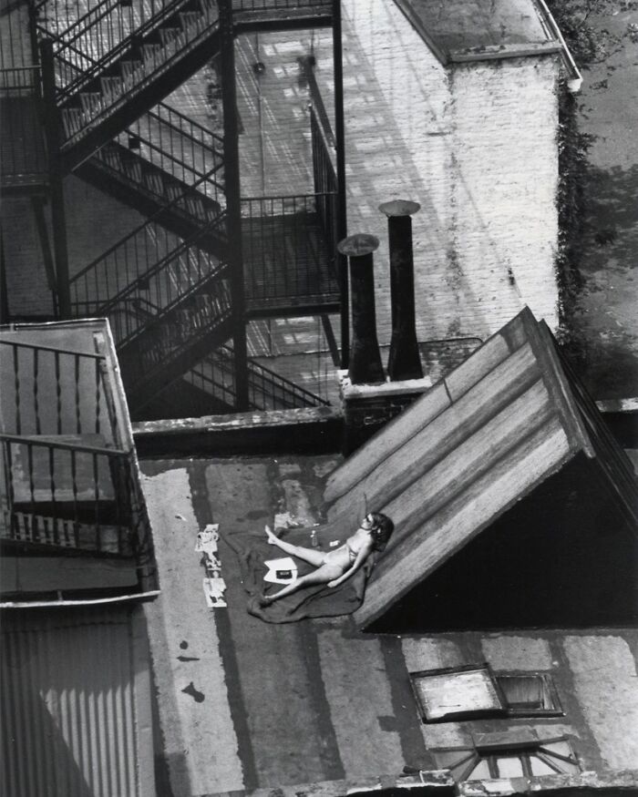 Black and white historic photo of a woman sunbathing on a rooftop with old chimneys and fire escapes visible.