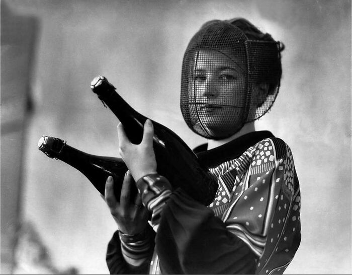 Young woman in vintage attire with a mesh face cover holding two old-style champagne bottles, history pics look confusing now.