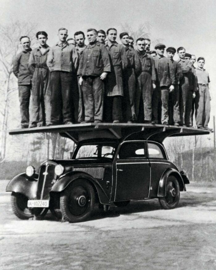 Group of men standing on a large platform supported by a vintage car in a confusing history photo from old times.