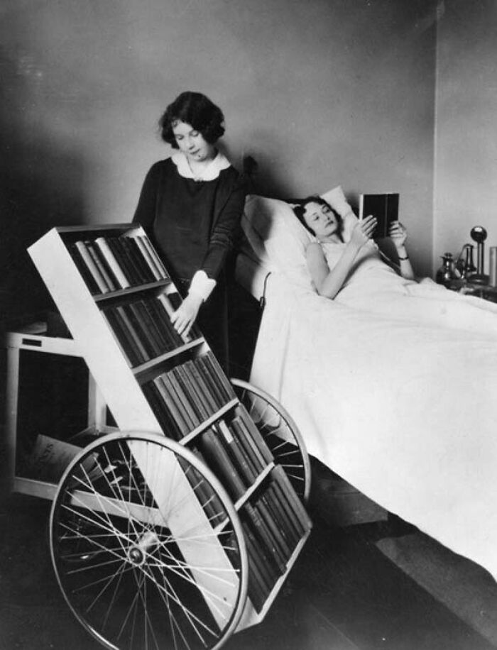 Woman using a vintage mobile book cart to read to a patient in bed in a historical photo that looks confusing now.