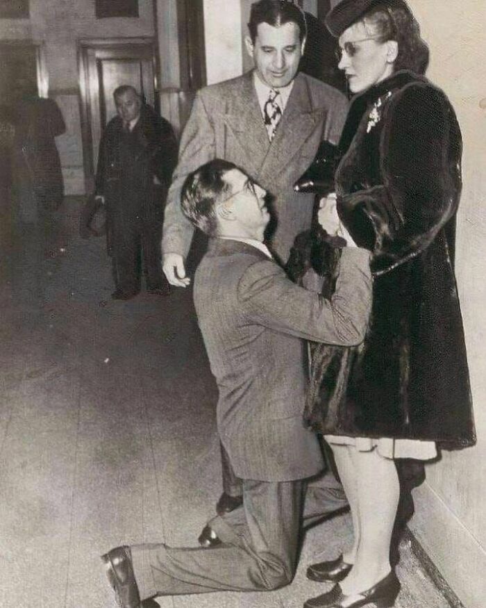 Black and white vintage photo showing a man kneeling while holding hands with a woman in historical clothing, strange history moment.