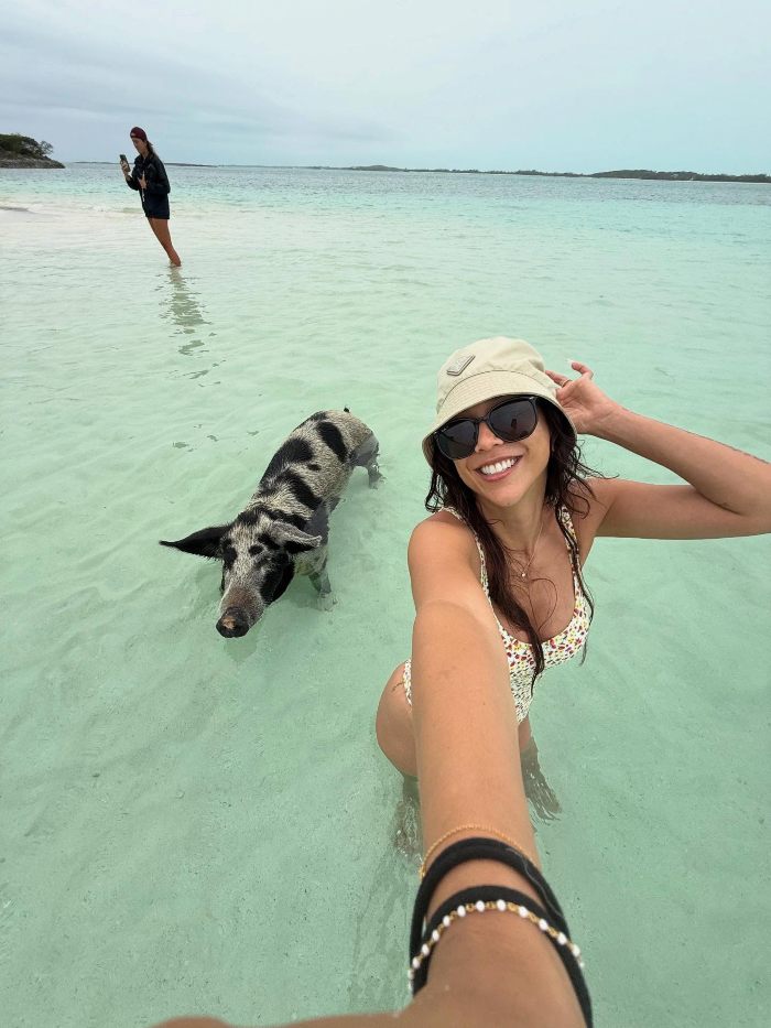 Aussie influencer in shallow water taking a selfie with a pig, wearing sunglasses and a hat, with another person far behind.