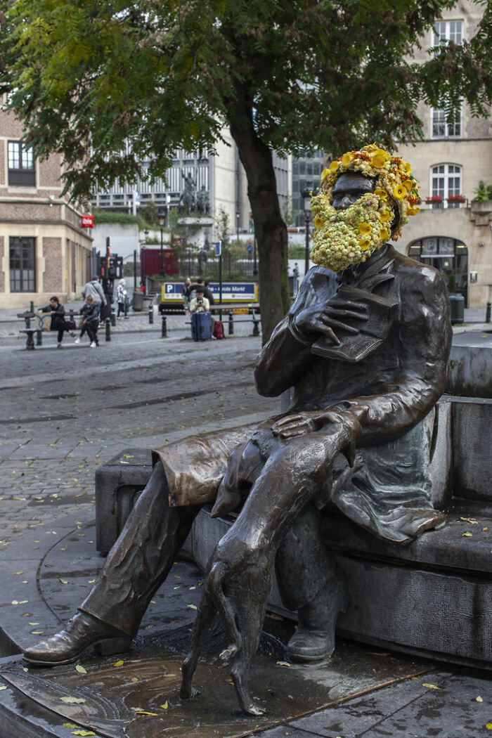 City statue dressed in flowers with floral beard and hair, sitting on a bench with a dog in an urban setting.