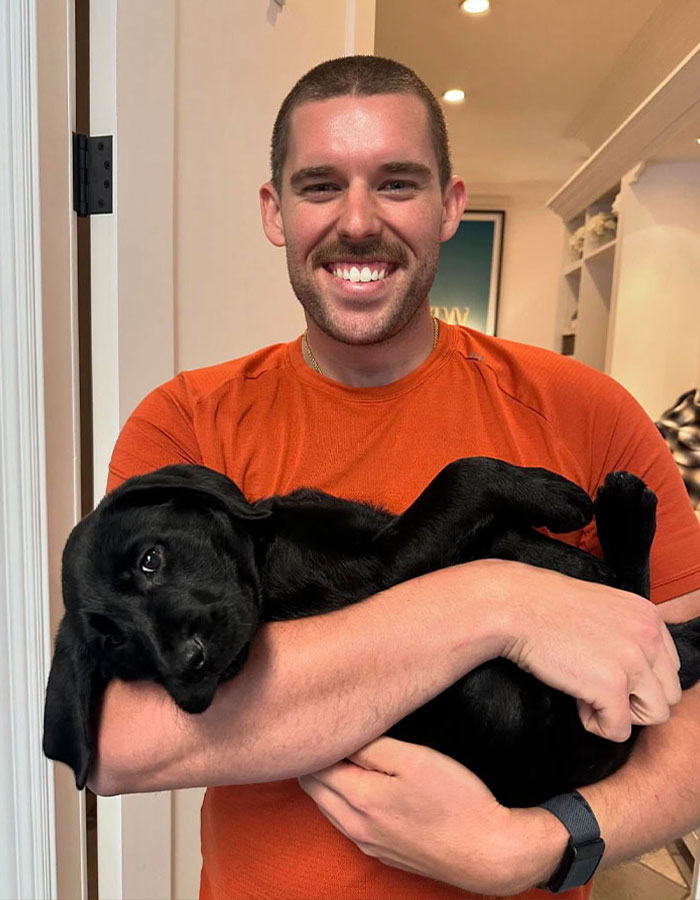 Christopher Schwarzenegger smiling, wearing orange shirt and holding a black dog indoors, showing dramatic weight loss. Christopher Schwarzenegger smiling, wearing orange shirt and holding a black dog indoors, showing dramatic weight loss.