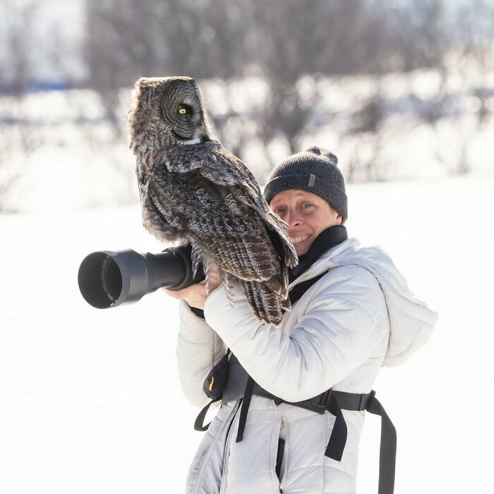 Wildlife photographer smiling with a large owl perched on camera lens during a snowy outdoor photo shoot.