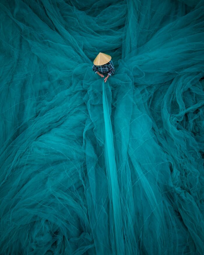 Person wearing a conical hat working with expansive blue nets in a stunning color photography shot.