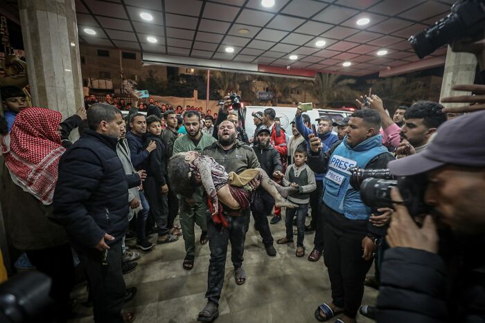 Crowd surrounded by press as man carries injured child in a powerful color photography image from award winners.