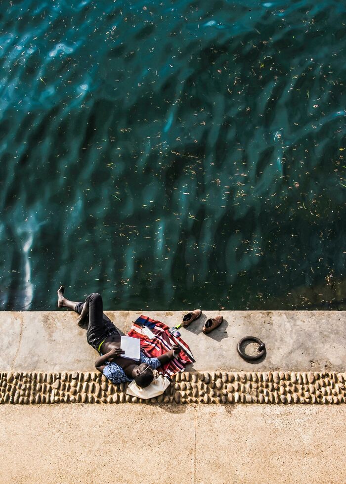 Man lying on riverside reading, with shoes and blanket nearby, showcasing stunning color photography composition.