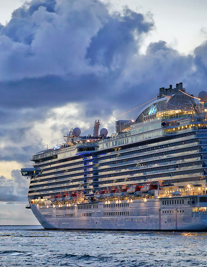 Cruise passenger standing onboard a large illuminated ship at dusk with cloudy sky in the background.