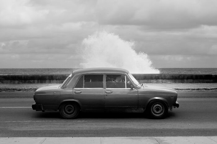 Black and white cinematic photo by Adriana Ferrarese showing a vintage car driving by the ocean with waves crashing in the background