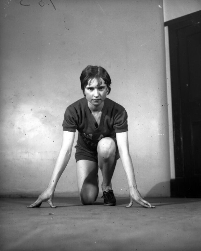 Black and white photo of a young woman in vintage athletic clothing in a starting runner's pose indoors, history looks confusing now