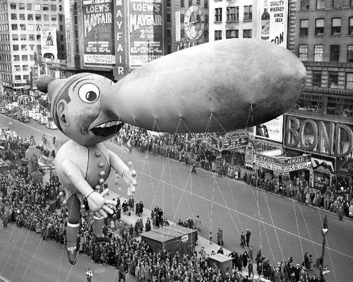 Giant vintage cartoon balloon floats over crowded city street during historical parade with confusing old photos.