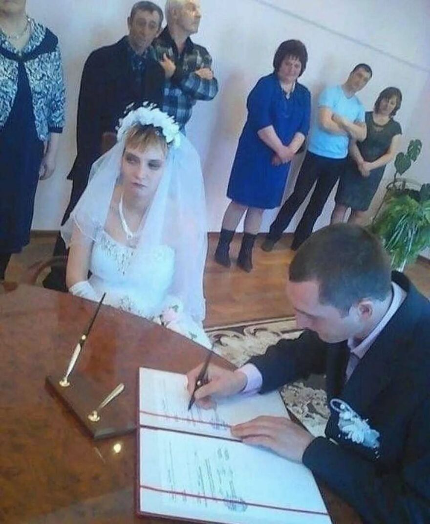 Bride looking unimpressed while groom signs documents during bizarre yet hilarious wedding ceremony with guests watching