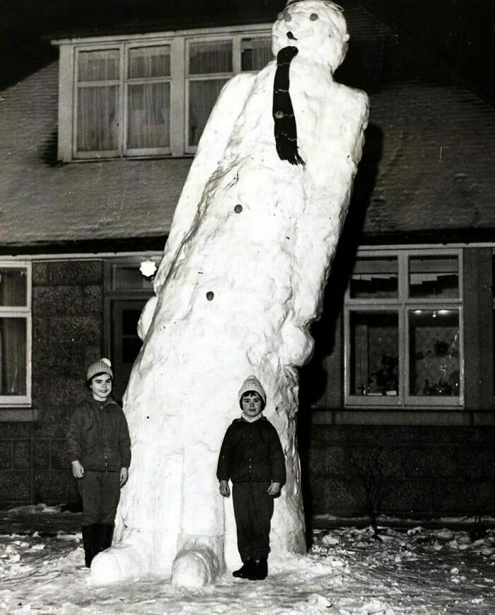 Two children standing next to a giant, unusually shaped snowman in a snowy yard, vintage confusing history photo.