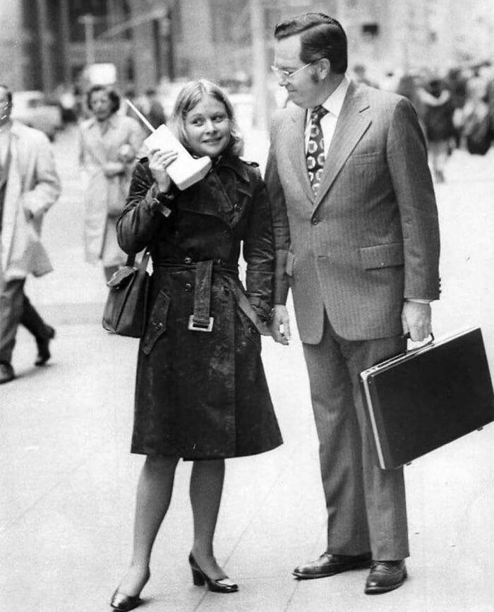 Black and white photo of a woman using a large vintage mobile phone, with a man in a suit holding a briefcase nearby in history.