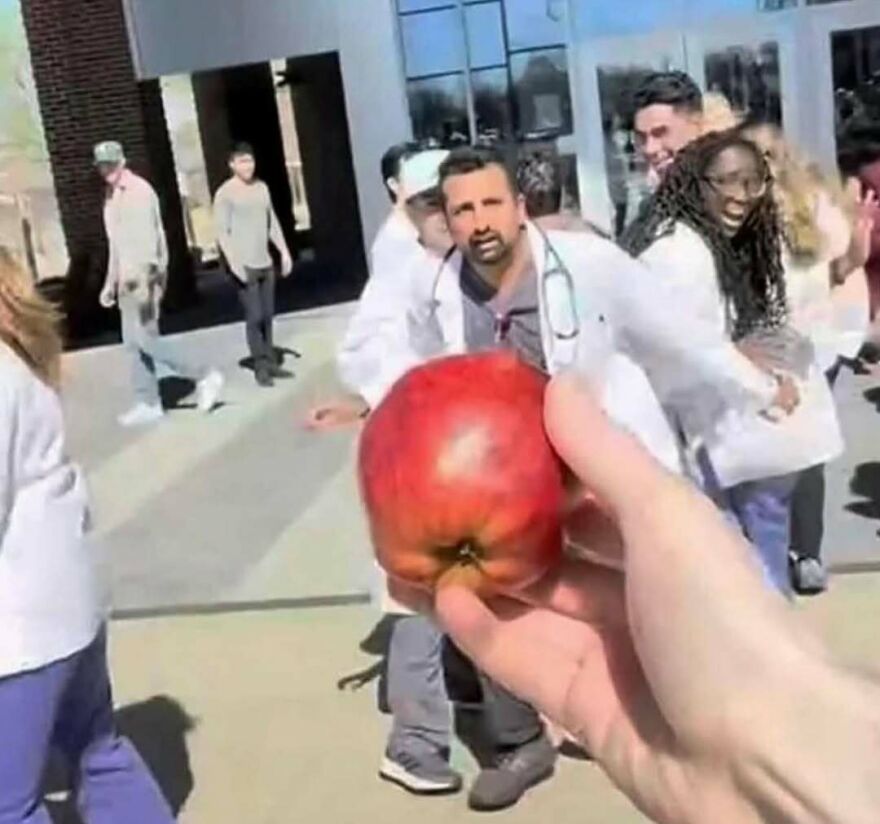 Hand holding an apple in the foreground with a group of people in white coats reacting, a bizarre yet hilarious pic.