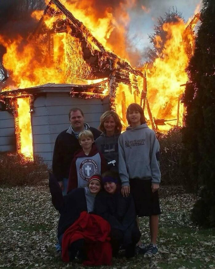 Family posing calmly in front of a large burning house, a bizarre image capturing an unusual moment in nature and animals context.